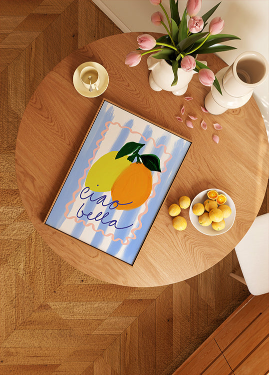 drewniany stół z oprawioną grafiką przedstawiającą dwie cytryny, miskę owoców i wazon z różowymi kwiatami.
